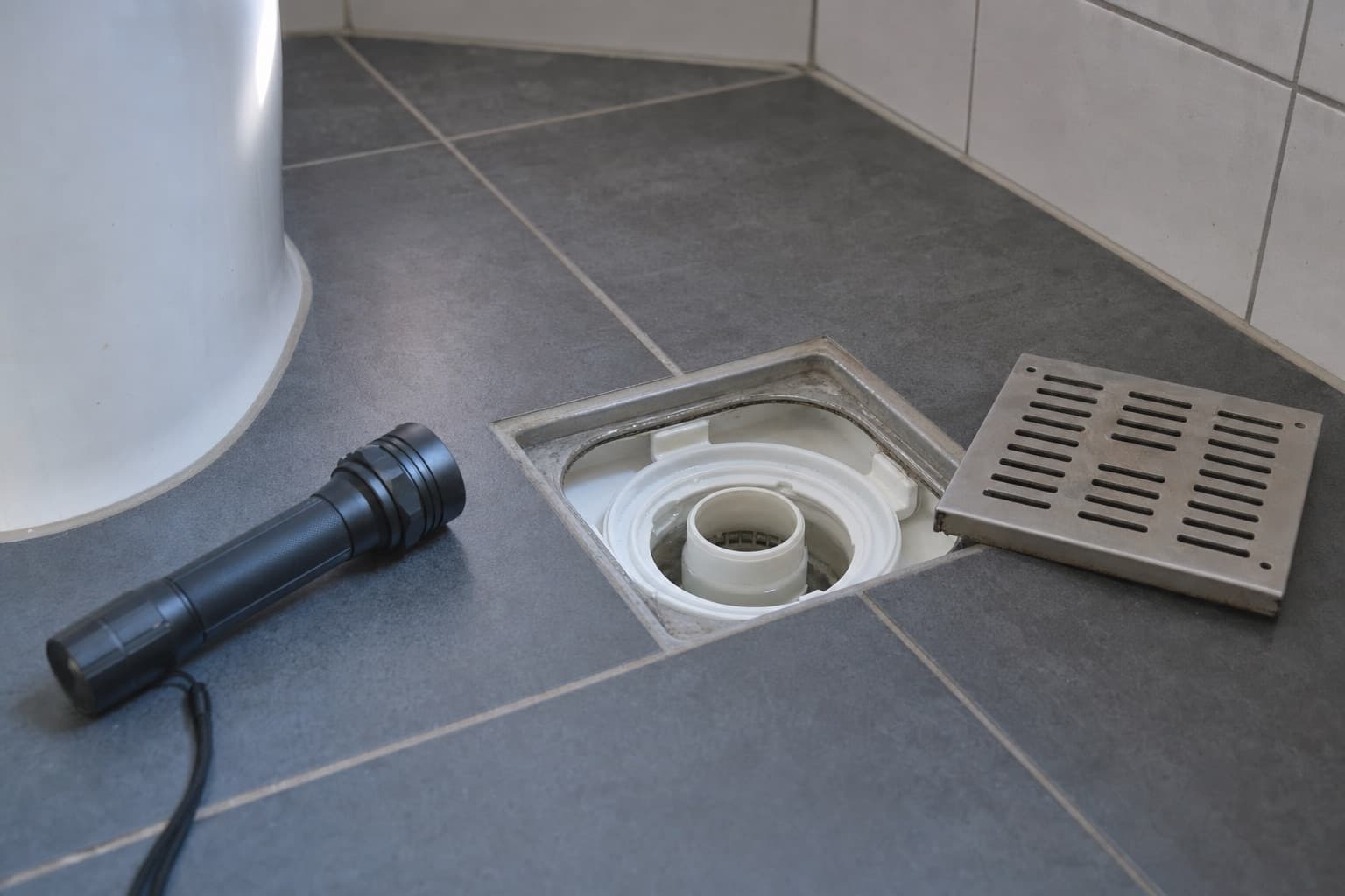 Le sol d’une salle de bains, la bonde de douche ouverte, le siphon visible et à sec, une lampe à côté